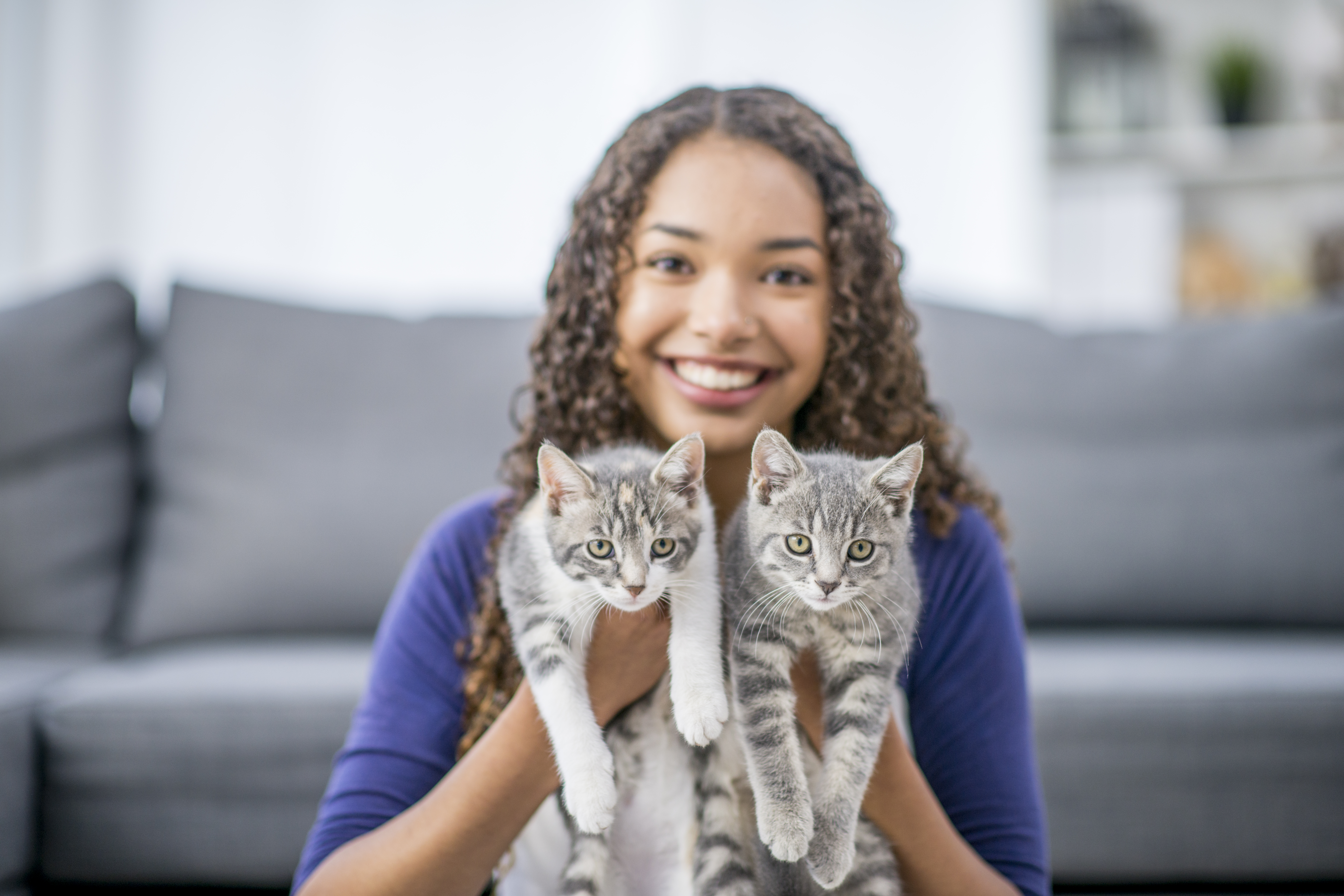 A girl of African descent is in her living room. She is holding up her two newly-adopted kittens, and smiling at the camera.